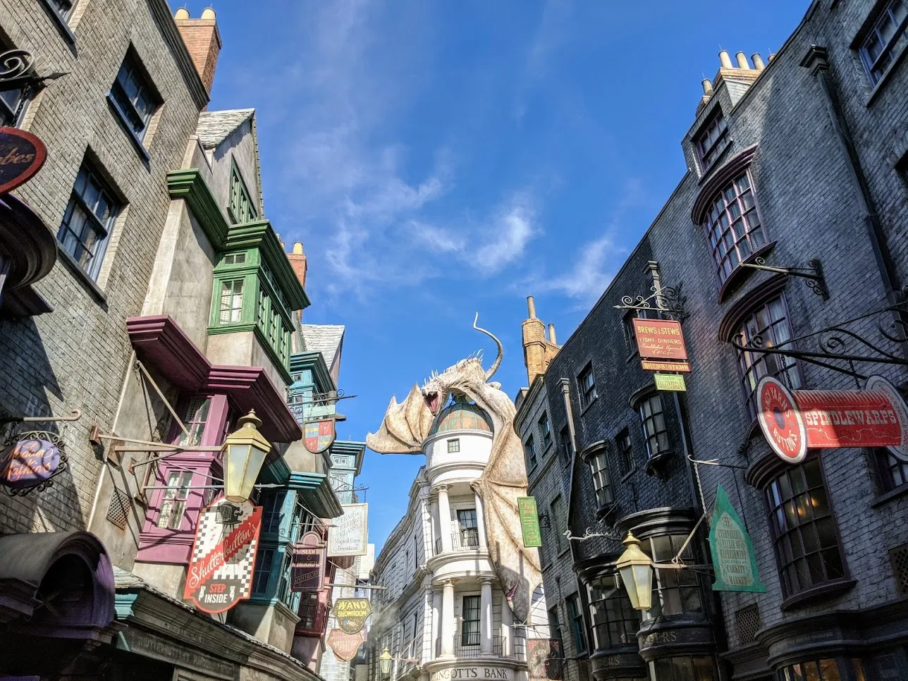 View of Diagon Alley at Universal Studios, featuring colorful shopfronts, hanging signs, and a fire-breathing dragon atop Gringotts Bank against a bright blue sky.