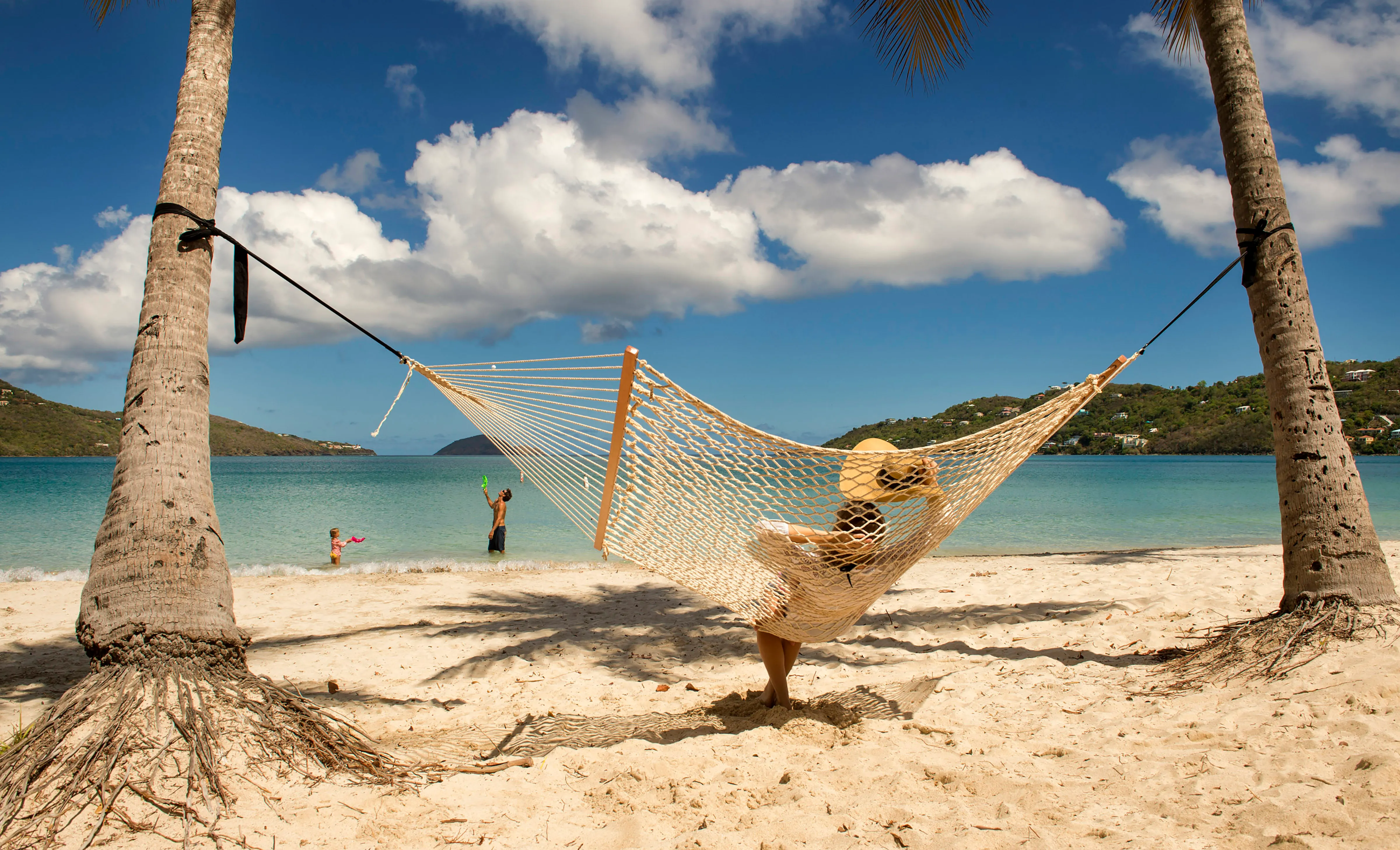 A woman in a hammock on the beach; in the background, a man and child are playing in the water near the shoreline.